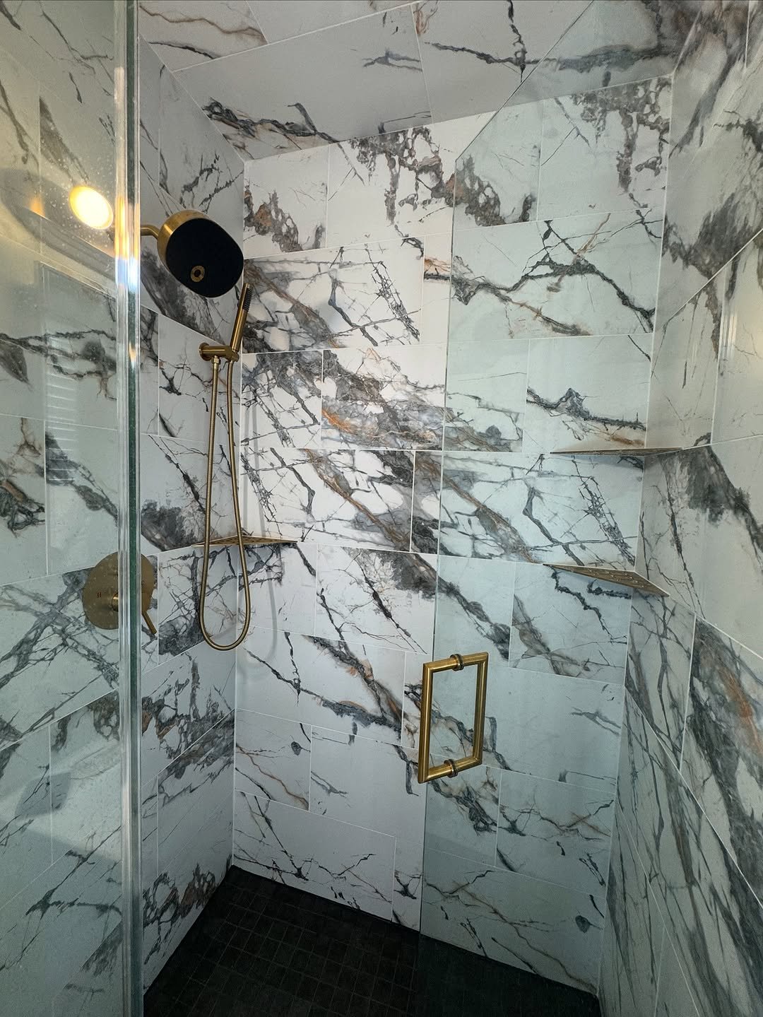 Walk-in shower clad floor to ceiling in dramatic veined marble-look tile with black and gold fixtures