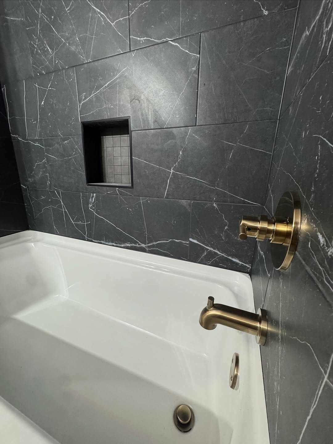 White freestanding tub against dark charcoal marble-look tile with a mosaic-lined niche and brass spout